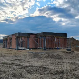 Surowa bryła domu w trakcie budowy, widoczna konstrukcja z czerwonej cegły i betonowy wieniec, wsparta metalowymi stemplami, na tle pochmurnego nieba.