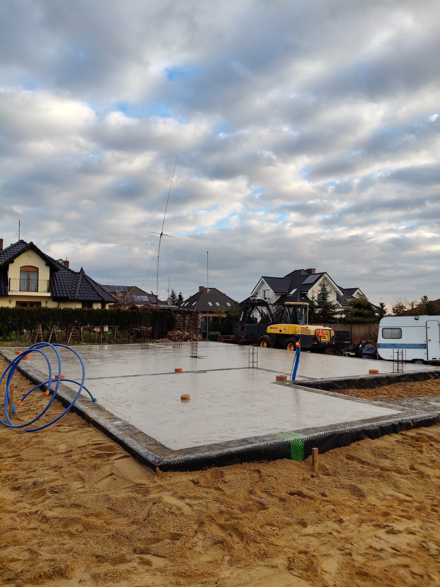 Wylany fundament pod budynek jednorodzinny z wystającymi prętami zbrojeniowymi, widoczny żółty traktor budowlany i przyczepa kempingowa w tle, pochmurne niebo.