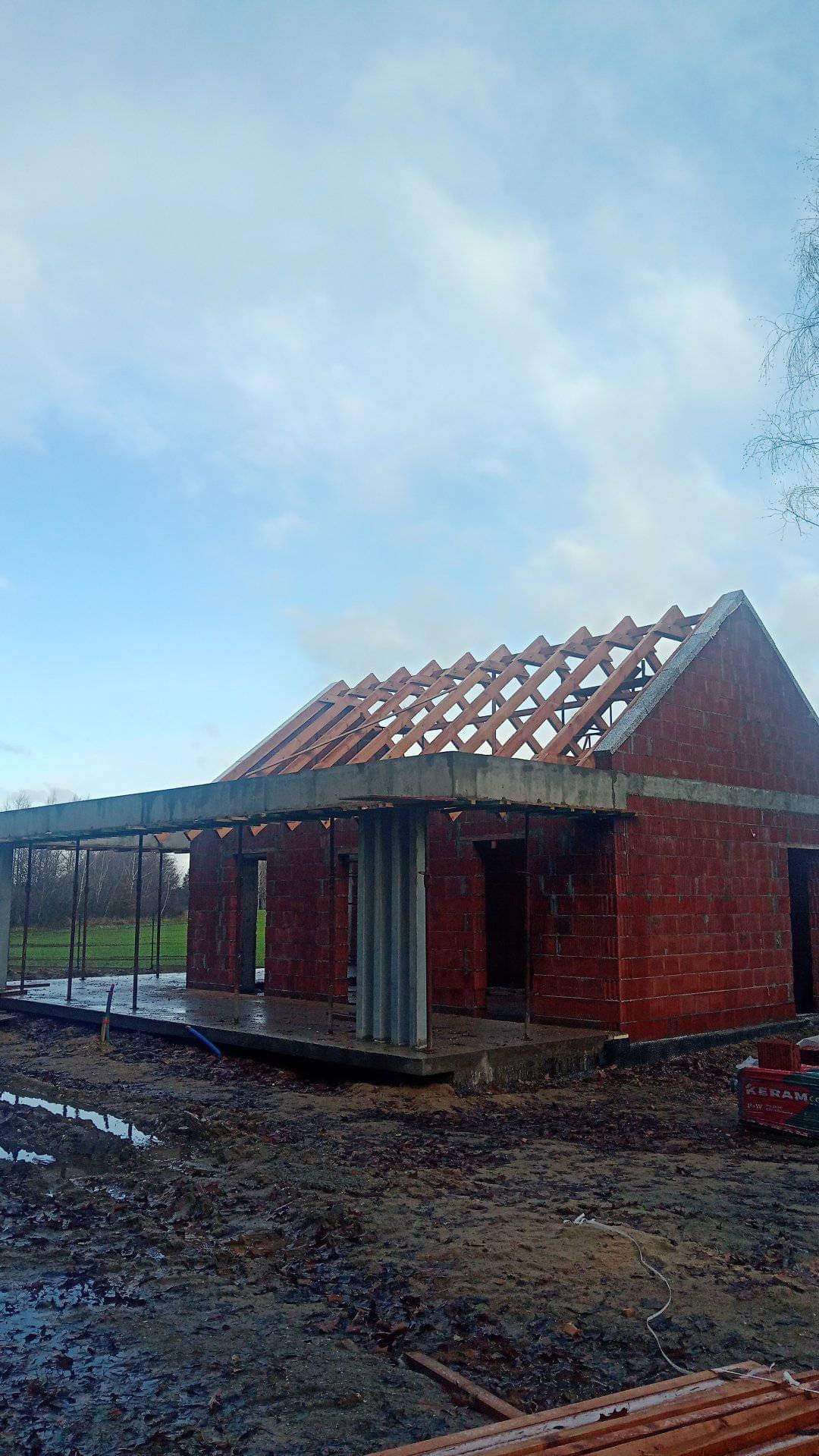 Surowy stan budowy domu jednorodzinnego z czerwonej cegły, widoczne drewniane krokwie dachu, betonowy wieniec i stalowe podpory, teren wokół placu budowy błotnisty.