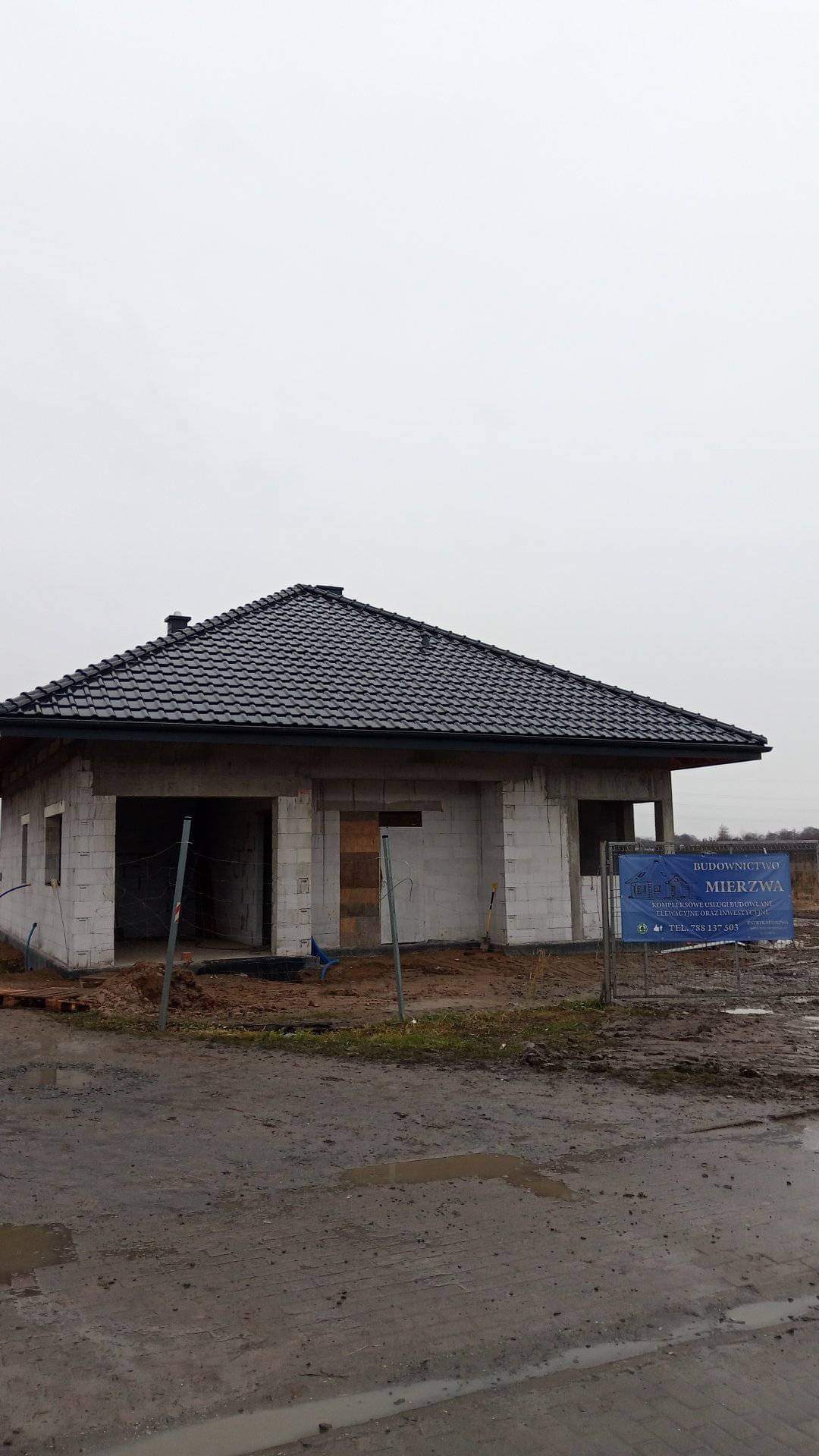 Surowy dom jednorodzinny z czarnym dachem z dachówki ceramicznej, w stanie deweloperskim, z widocznymi bloczkami betonu komórkowego, na błotnistym terenie, z tablicą reklamową firmy budowlanej w tle.