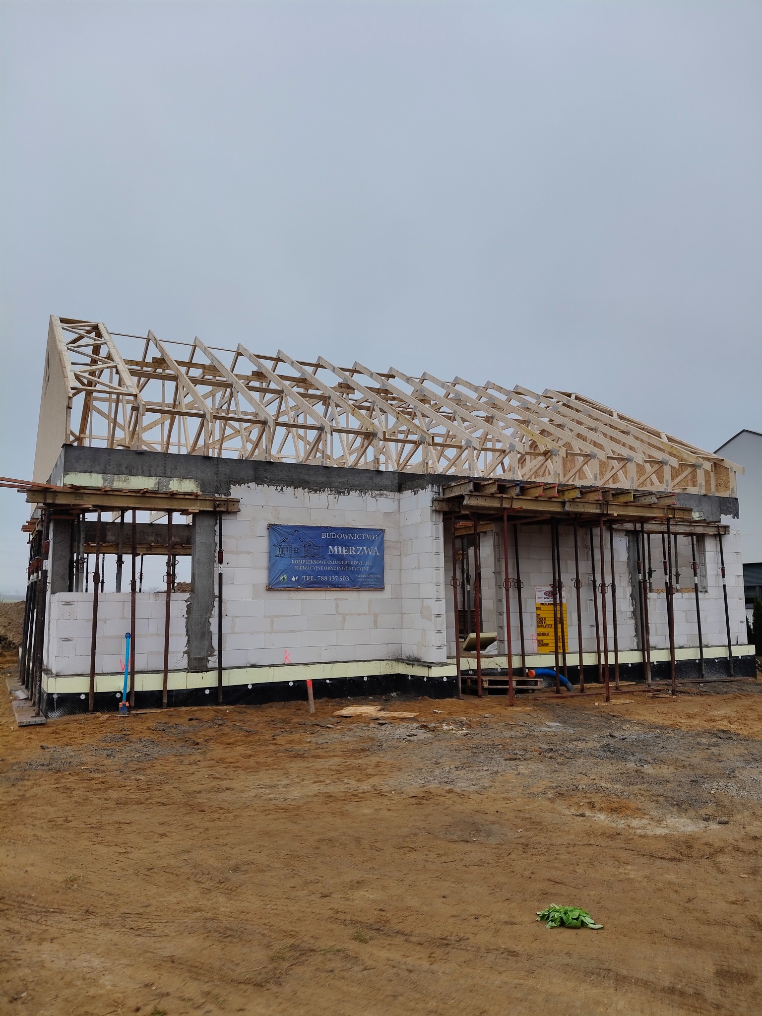 Surowy stan budynku jednorodzinnego w trakcie budowy: ściany z bloczków, drewniana konstrukcja dachu bez pokrycia, podpory stalowe, tablica informacyjna budowy na ścianie.