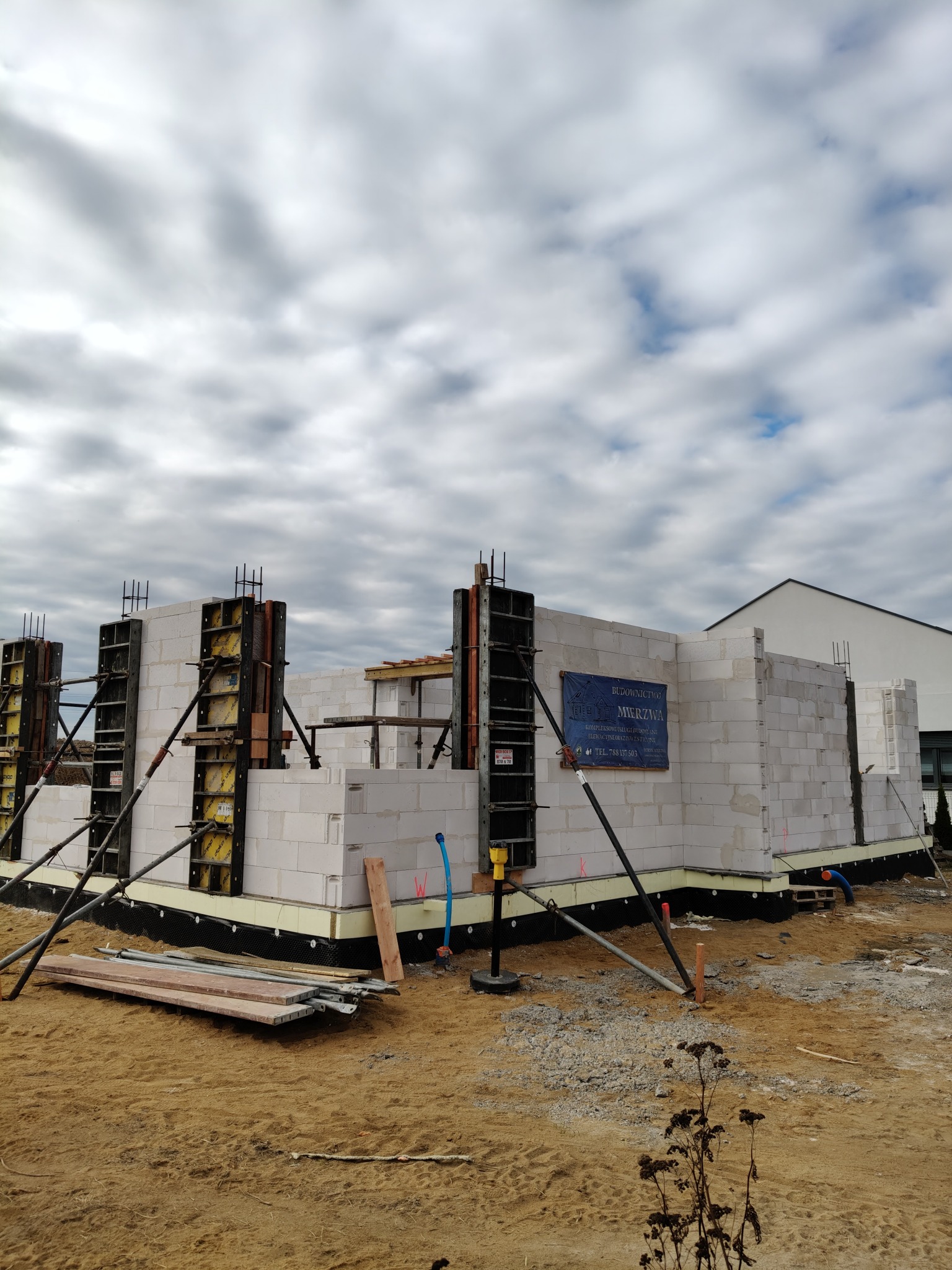 Wznoszenie ścian domu z bloczków betonowych, widoczne szalunki, izolacja fundamentów, banner reklamowy firmy budowlanej na ścianie, piaszczysty teren budowy.