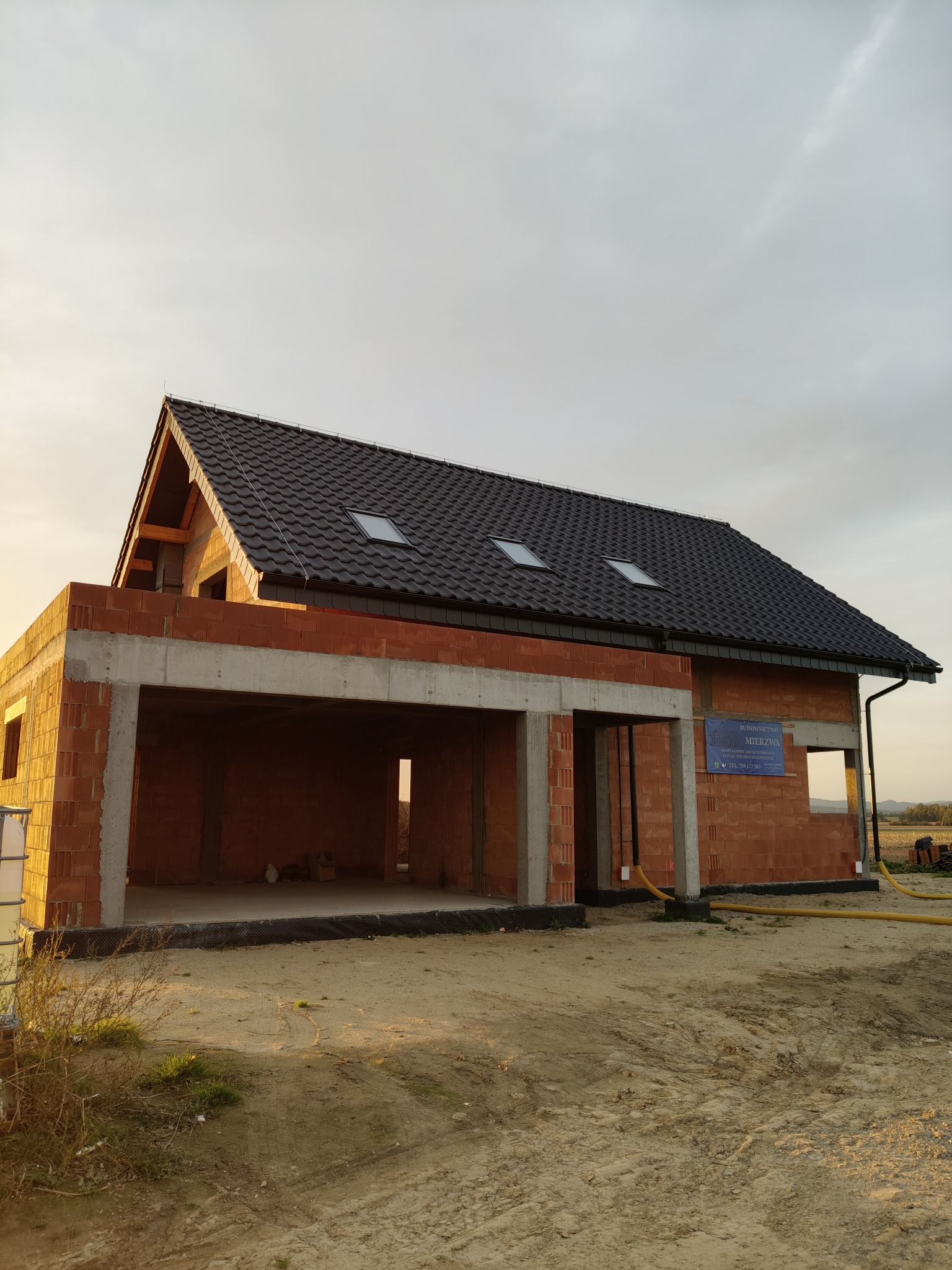 Surowy stan domu jednorodzinnego z garażem w trakcie budowy, widoczne ściany z czerwonej cegły, czarny dach z trzema oknami dachowymi, żółte węże na zewnątrz budynku.