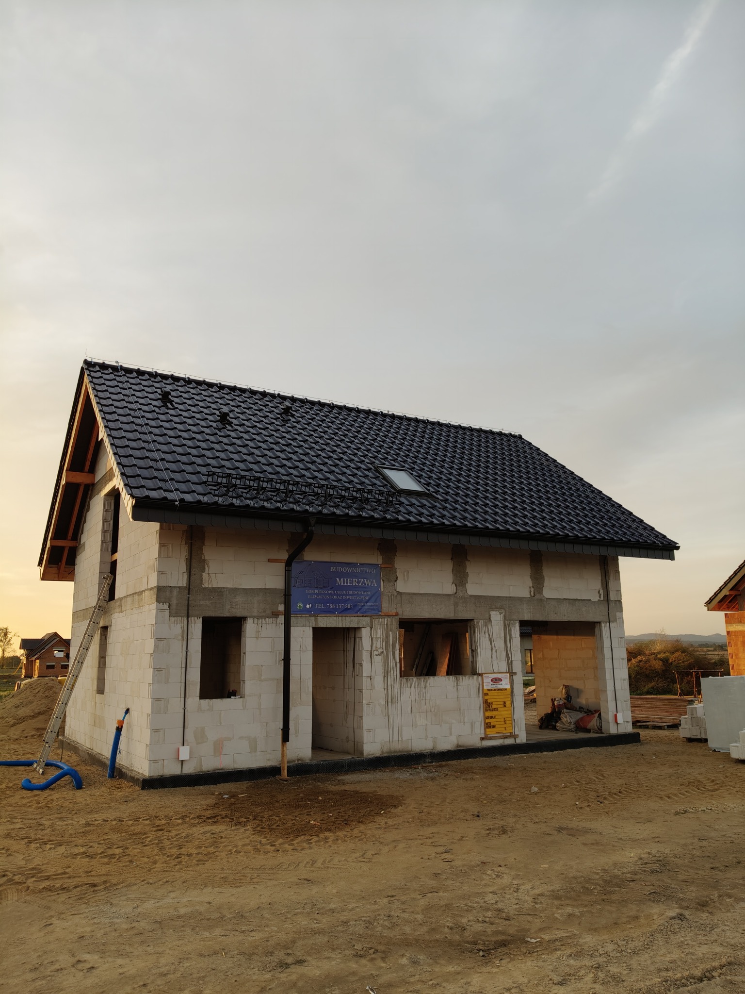 Surowy stan domu jednorodzinnego w trakcie budowy w miejscowości Biała, z widocznym czarnym dachem z dachówki, ścianami z białego bloczka i zamontowaną rynną. Na ścianie wisi tablica informacyjna...