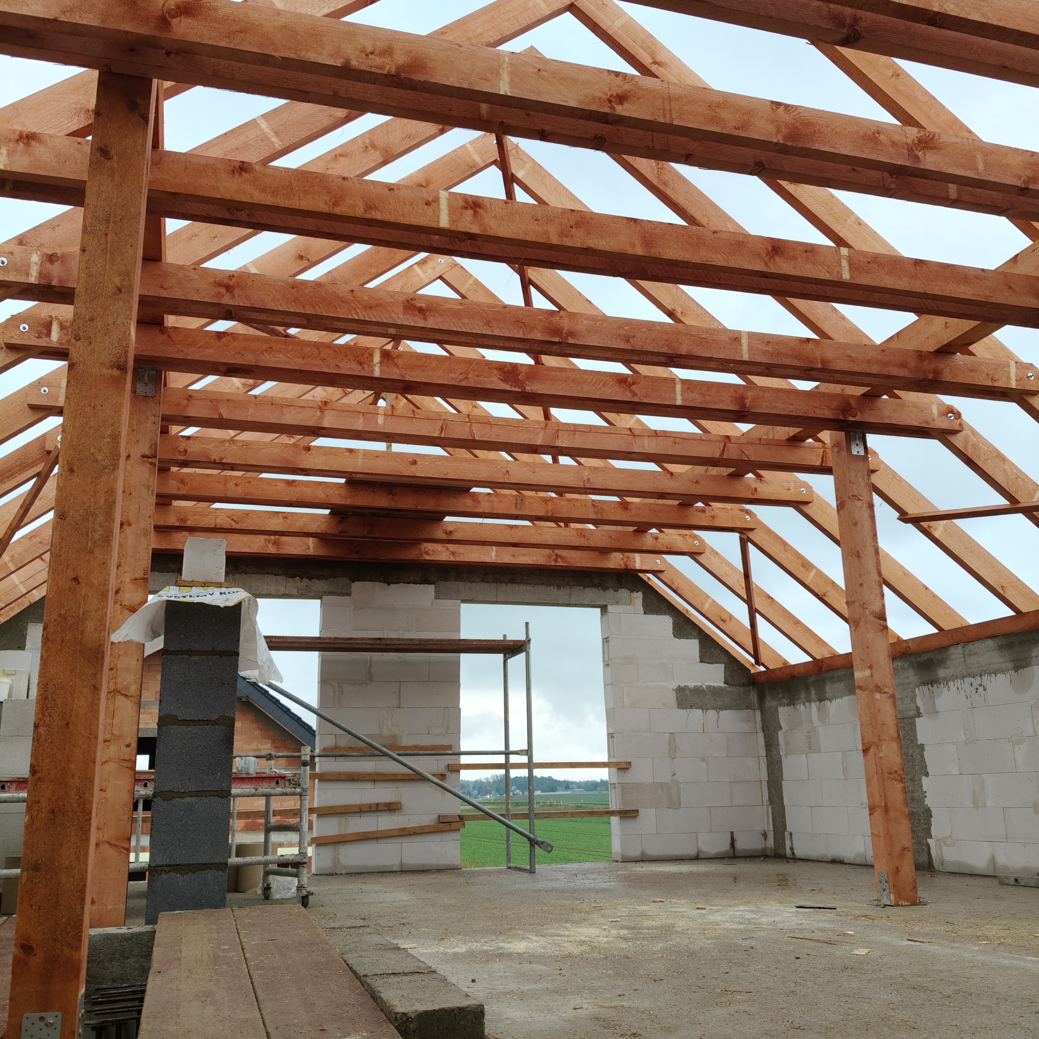 Konstrukcja drewniana dachu w trakcie budowy domu z bloczków betonowych, widoczne elementy więźby dachowej i ściany w surowym stanie.