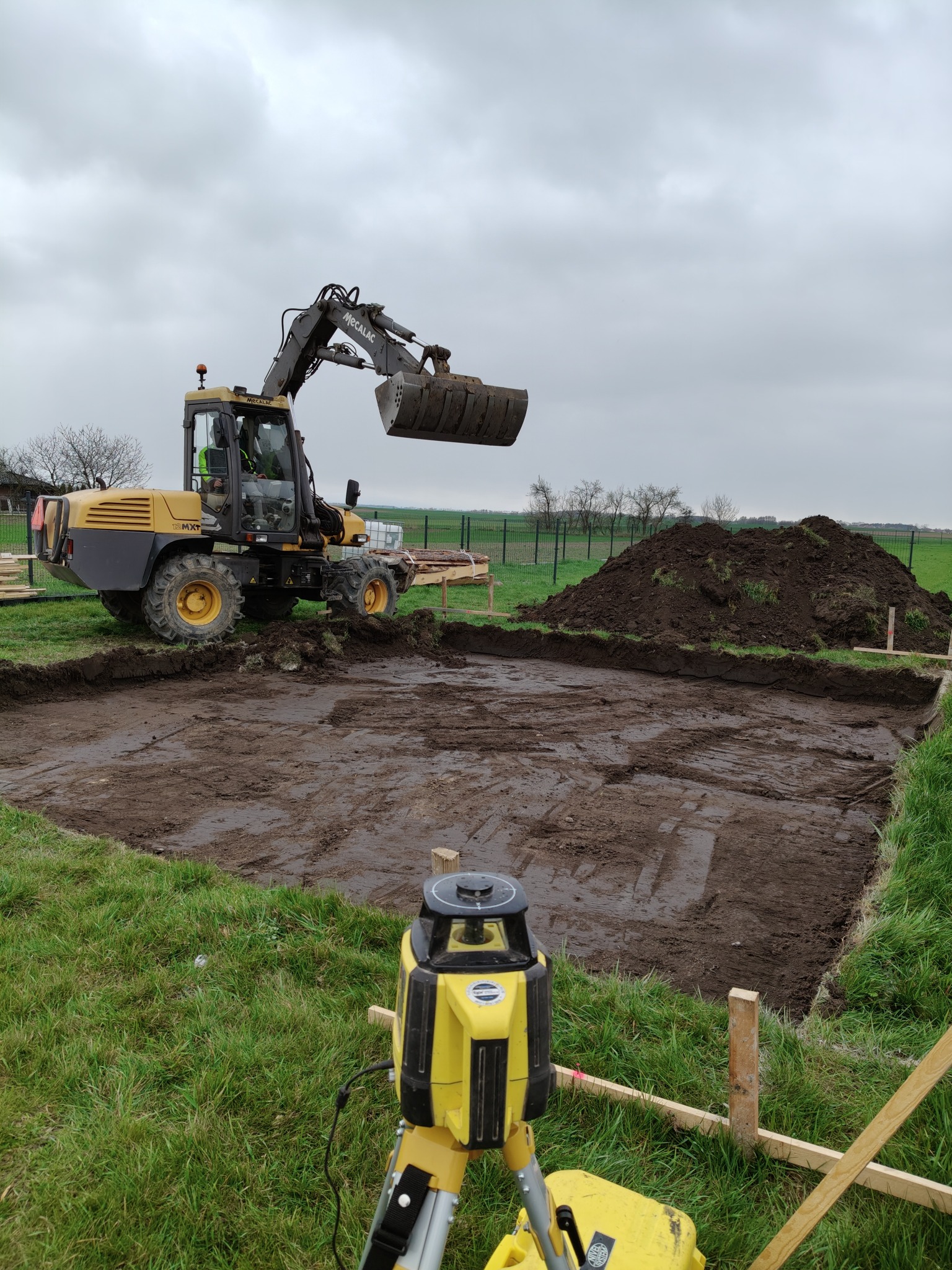 Żółta koparka Mecalac podczas prac ziemnych na budowie domu. W tle hałda ziemi i ogrodzenie, a na pierwszym planie niwelator laserowy.