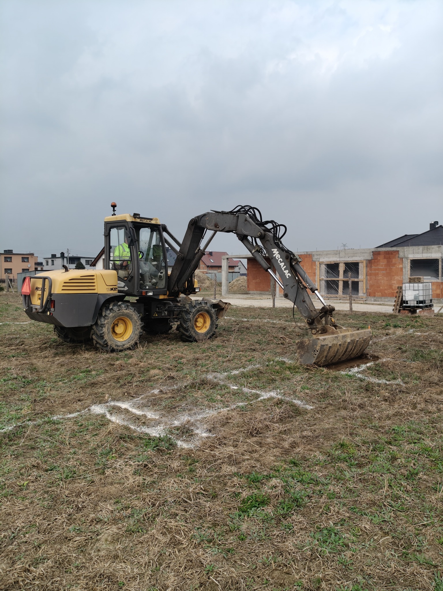 Żółto-szara koparka Mecalac na placu budowy, w tle budynek w trakcie wznoszenia z czerwonej cegły. Na ziemi widoczne białe oznaczenia fundamentów.