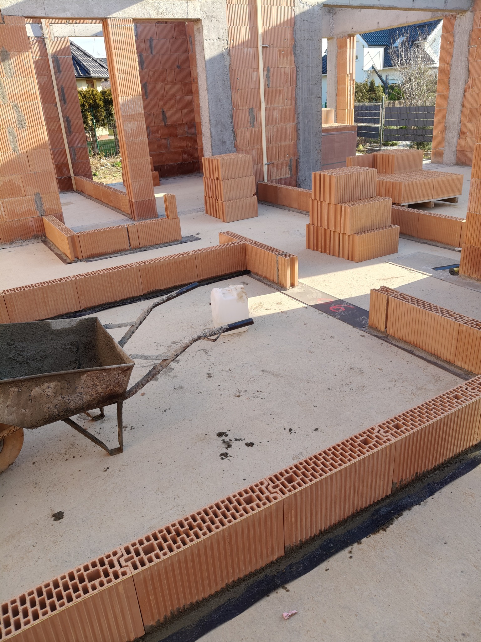 Budowa domu z pustaków ceramicznych: ściany wzniesione do pewnej wysokości, widoczna taczka z zaprawą murarską, pustaki ułożone na podłodze. Etap wznoszenia ścian.