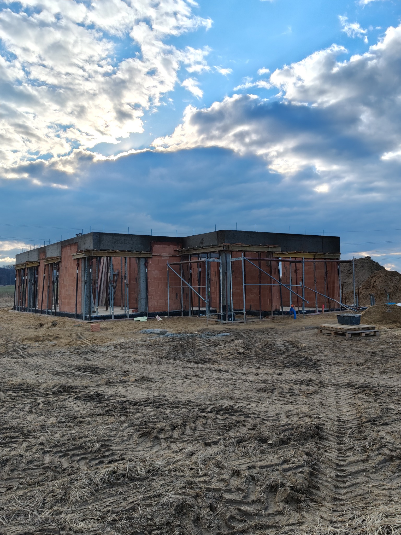 Surowa bryła domu w trakcie budowy, widoczna konstrukcja z czerwonej cegły i betonowy wieniec, wsparta metalowymi stemplami, na tle pochmurnego nieba.