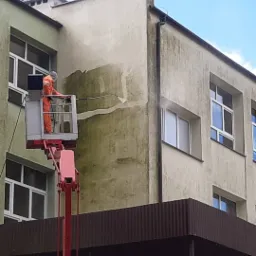 Pracownik w pomarańczowym kombinezonie, stojący na podnośniku koszowym, czyści elewację budynku za pomocą myjki ciśnieniowej, usuwając zielony nalot.