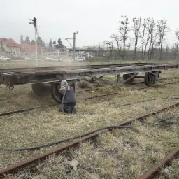 Pracownik w kombinezonie ochronnym czyści metalowy wagon kolejowy z rdzy i farby za pomocą strumienia ściernego na zewnątrz, na tle torów kolejowych i zabudowań.