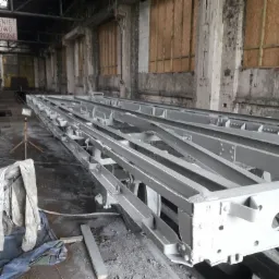 Szara, pomalowana proszkowo konstrukcja stalowa wagonu kolejowego w hali przemysłowej w Nowym Tomyślu, widoczne ślady wcześniejszej korozji na ścianach budynku.