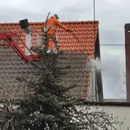 Pracownik w pomarańczowym kombinezonie na podnośniku koszowym czyści dach z czerwonej dachówki strumieniem wody pod ciśnieniem, widoczny świerk na pierwszym planie.