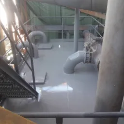 Wnętrze przemysłowe z metalowymi rurami, schodami i świeżo pomalowaną szarą podłogą, widoczne ślady mokrej farby, oświetlone przez naturalne światło wpadające z góry.