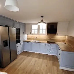 Jasna kuchnia w odcieniach błękitu i bieli z drewnianym blatem i panelem ściennym, oświetlona wiszącą lampą w stylu industrialnym.