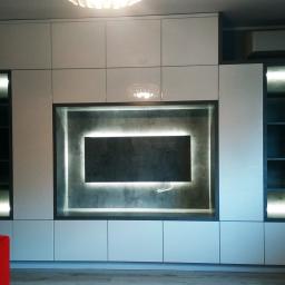 PIETRAS MEBLE - Nowoczesna zabudowa ściany z białymi szafkami, podświetlanymi półkami i wnęką na telewizor z betonowym tłem i ledowym podświetleniem.