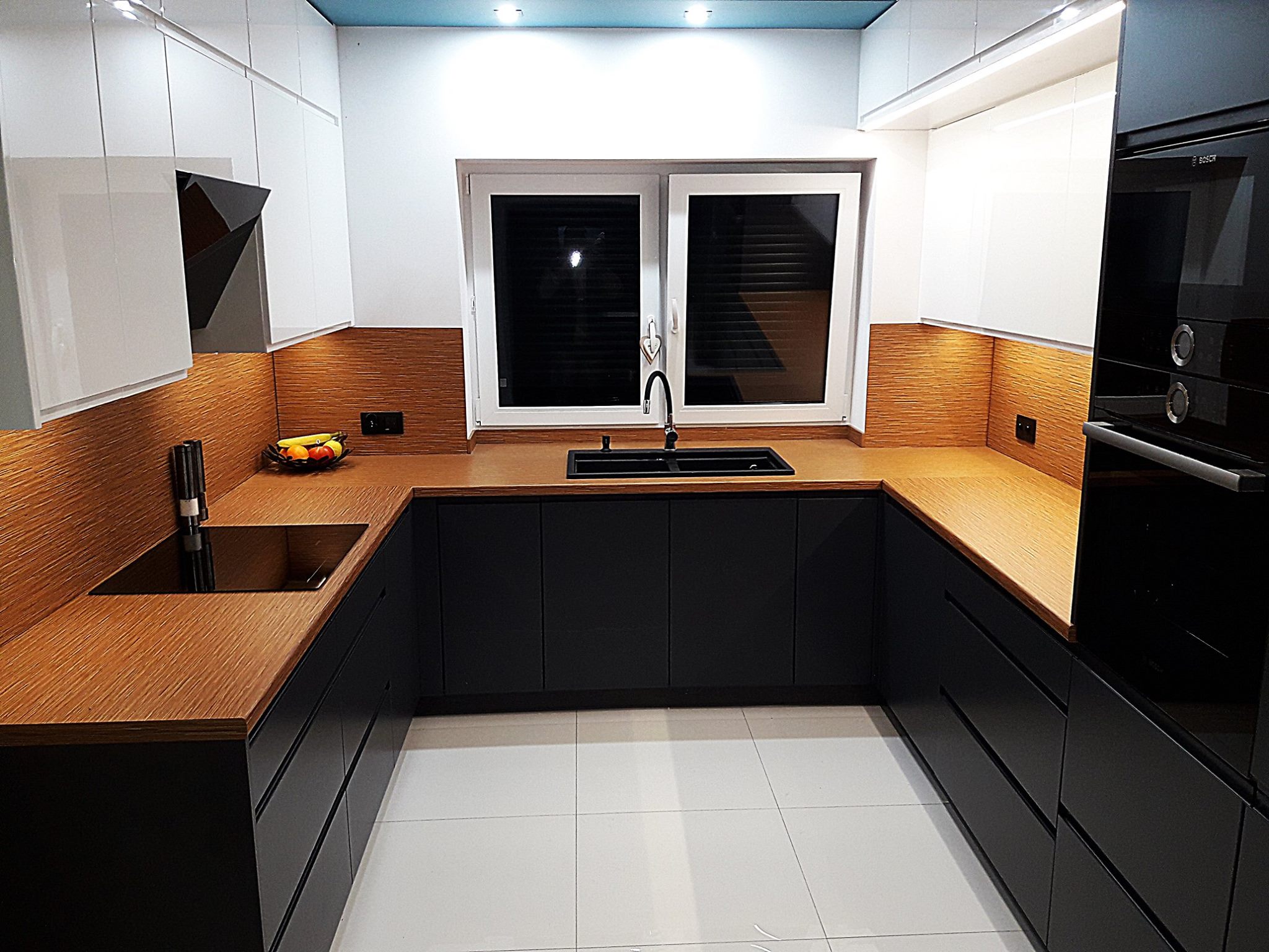 Nowoczesna kuchnia w odcieniach bieli i czerni z drewnianym blatem, czarny zlew i bateria, białe płytki podłogowe, oświetlenie podszafkowe LED.