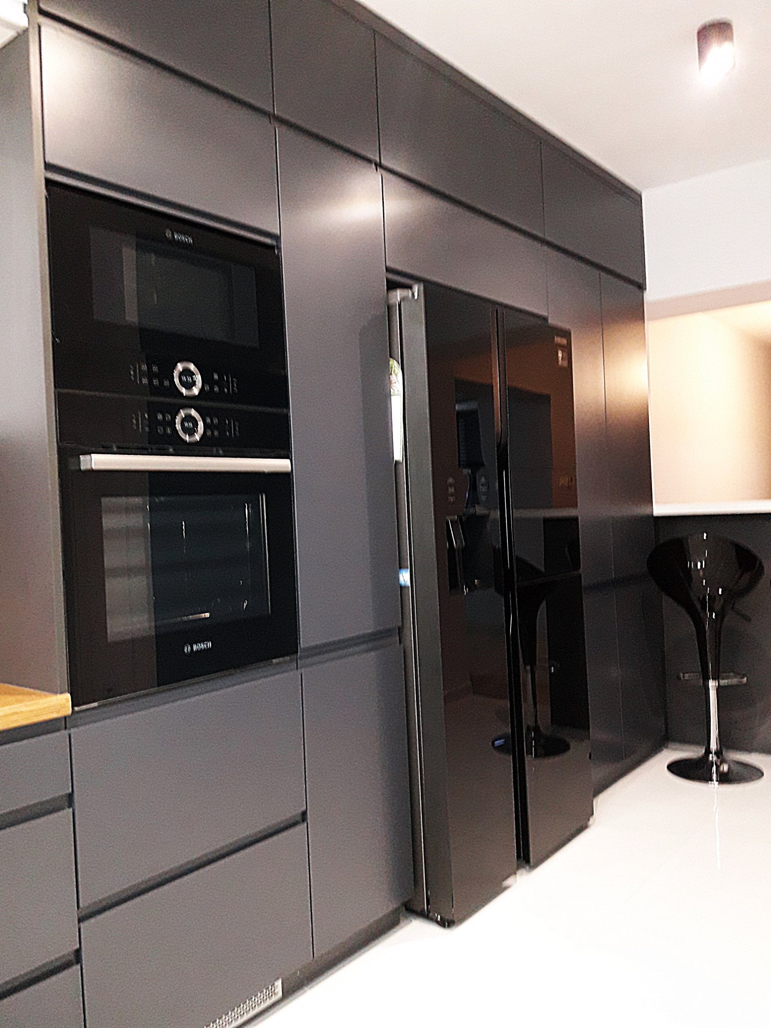 Nowoczesna kuchnia w odcieniach szarości z zabudowanymi urządzeniami marki Bosch i dużą lodówką side-by-side, uzupełniona o czarny hoker.