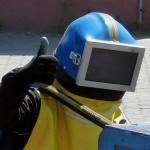 Spawacz w niebieskim hełmie z wizjerem, żółtej kamizelce i czarnych rękawicach unosi kciuk w górę. Widoczny fragment metalowej konstrukcji i szary mur w tle.