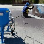 Pracownik w stroju ochronnym piaskuje metalową skrzynię na zewnątrz, obok stoi niebieski piaskarka. Widoczne cienie i przewody. Jasne oświetlenie.