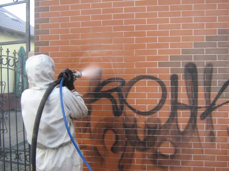 Czyszczenie ceglanej ściany z graffiti za pomocą myjki ciśnieniowej. Pracownik w kombinezonie ochronnym i masce. Widoczny fragment metalowego ogrodzenia.