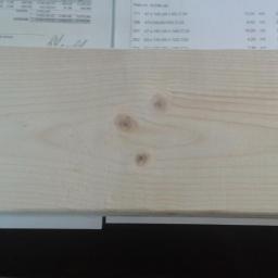 WealthBay S.A - Jasna deska z widocznymi słojami i trzema sękami, leżąca na tle faktury dokumentu z częściowo widocznymi danymi i czarnym blatem. Detal tarcicy budowlanej.