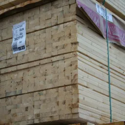 Stos tarcicy budowlanej, widoczne etykiety z numerami seryjnymi i logo producenta. Drewno ułożone równo, zabezpieczone folią i taśmą. Jasne oświetlenie.