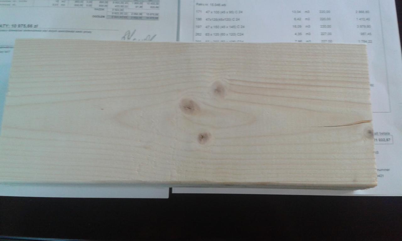 Jasna deska z widocznymi słojami i trzema sękami, leżąca na tle faktury dokumentu z częściowo widocznymi danymi i czarnym blatem. Detal tarcicy budowlanej.