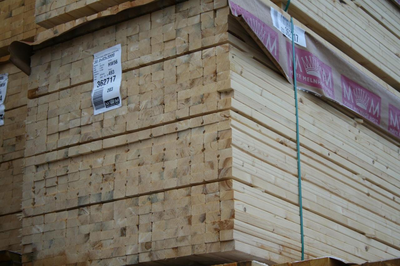 Stos tarcicy budowlanej, widoczne etykiety z numerami seryjnymi i logo producenta. Drewno ułożone równo, zabezpieczone folią i taśmą. Jasne oświetlenie.