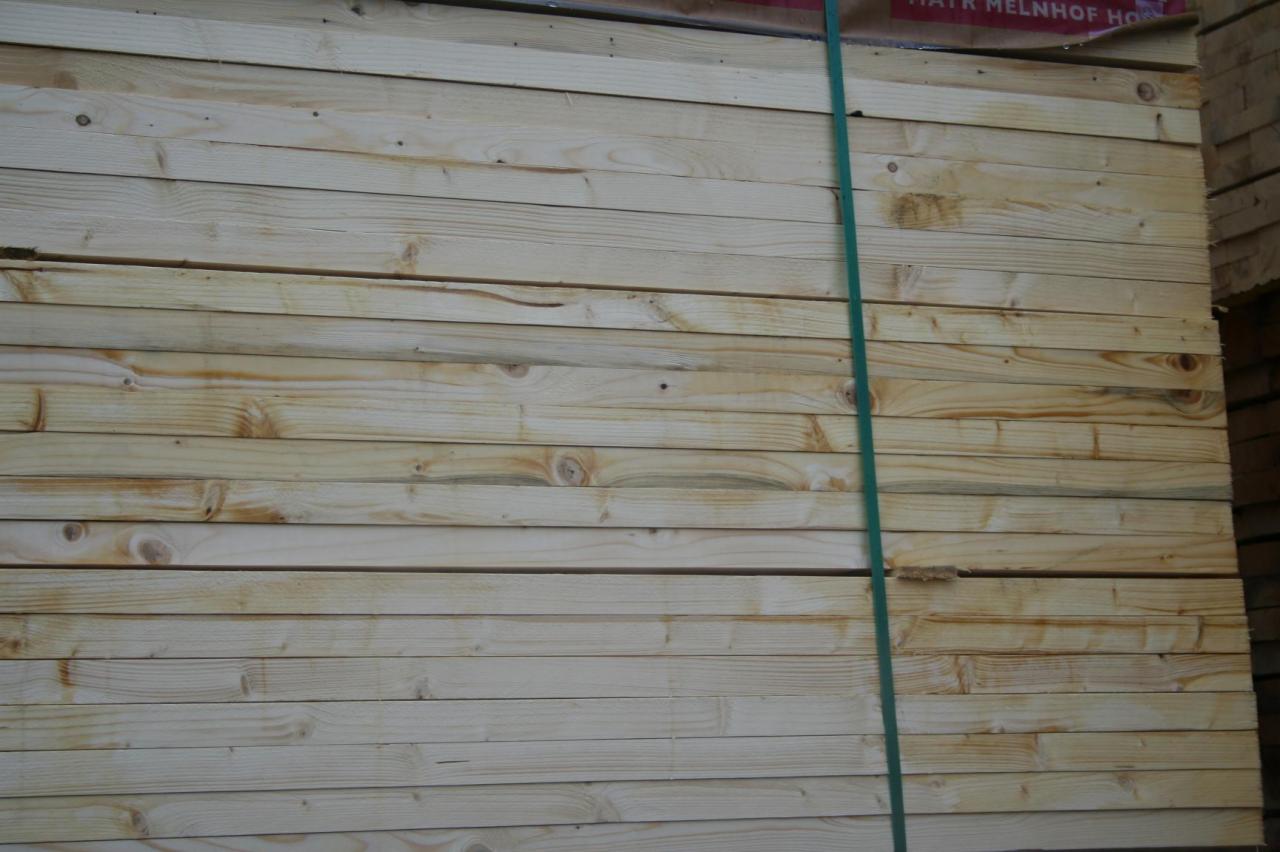 Stos jasnych desek tarcicy budowlanej, spiętych zieloną taśmą, z widocznym fragmentem etykiety producenta w górnej części kadru.