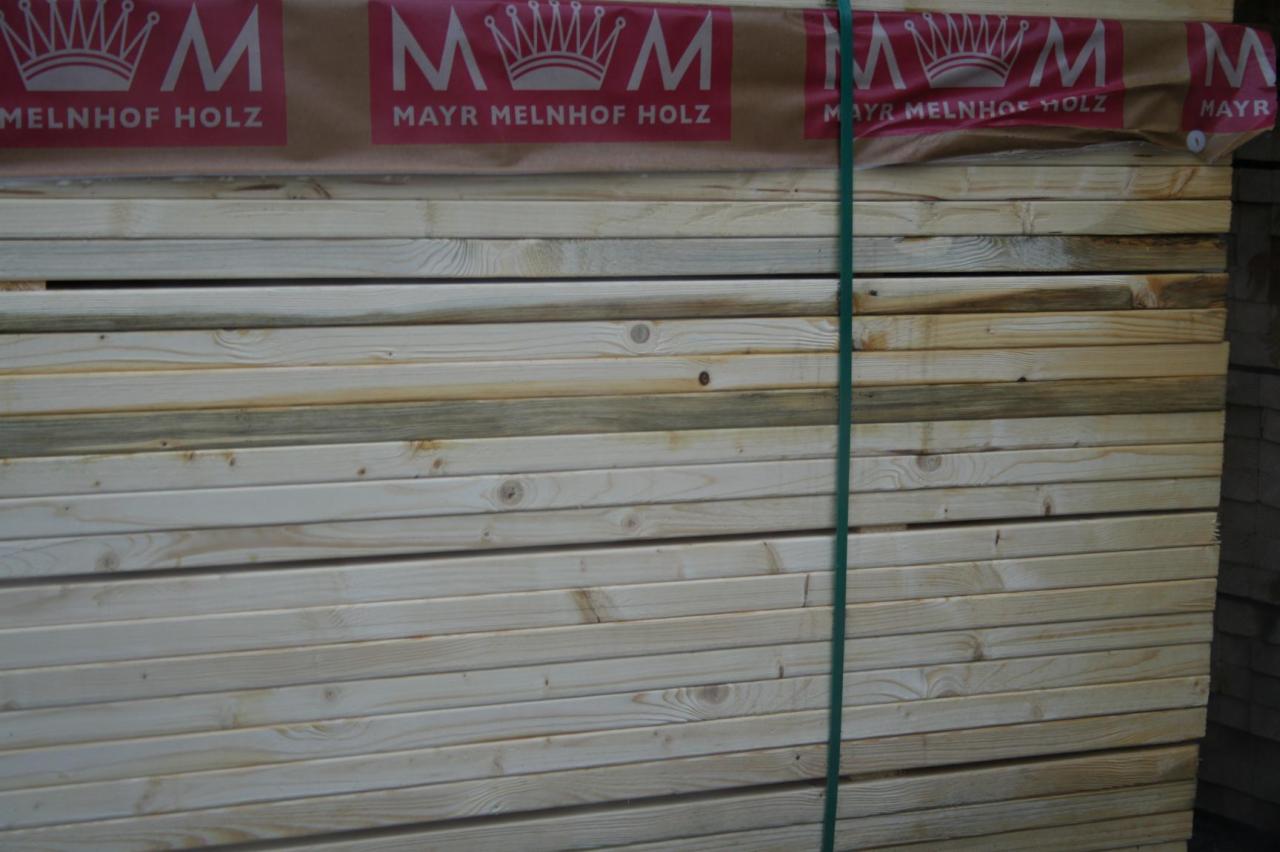 Stos tarcicy budowlanej związany zieloną taśmą, widoczny fragment logotypu producenta na opakowaniu. Jasne drewno z widocznymi słojami.