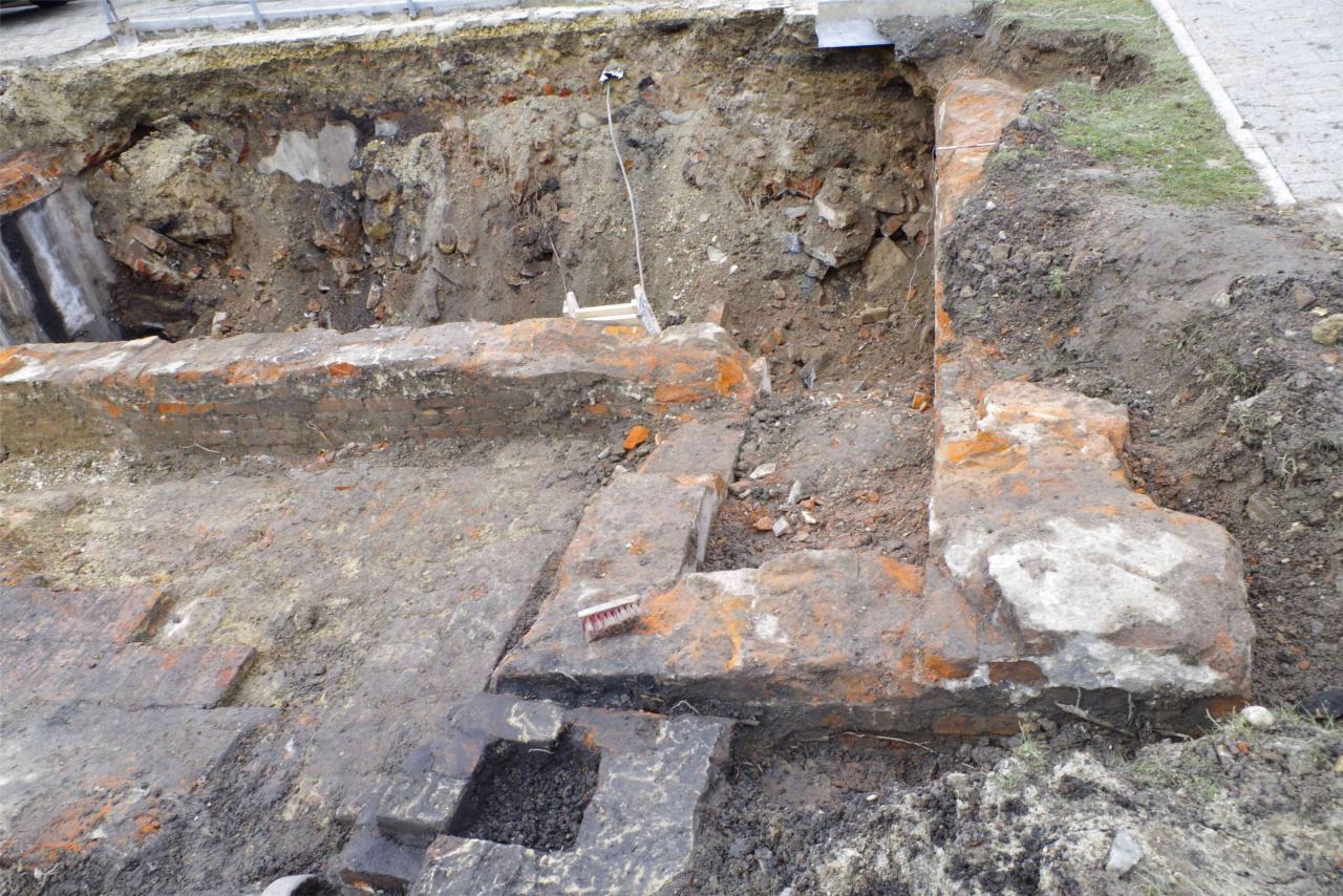 Odkopane fundamenty ceglane w ziemi, widoczne fragmenty muru i wykop, szczotka leżąca na murze. Prace ziemne podczas budowy lub remontu.