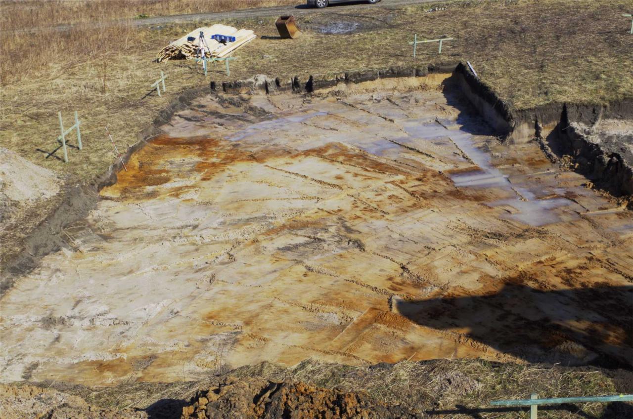 Wykonany wykop pod fundamenty domu z widocznymi śladami po pracy koparki. Na górze sterta desek i narzędzia pomiarowe. Widok z góry.