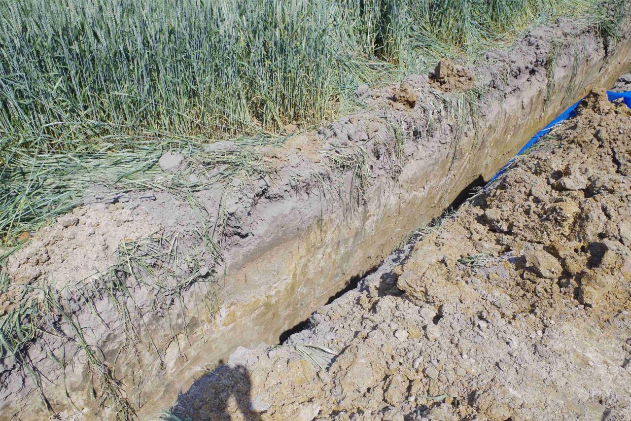 Wykop w ziemi z widoczną niebieską rurą, częściowo zasypany ziemią i trawą. Widoczny cień osoby w lewym dolnym rogu. Prace ziemne przy instalacji.