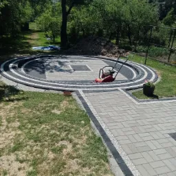 Okrągły plac z kostki brukowej w odcieniach szarości i czerni, otoczony zielenią, z zagęszczarką w centrum i ścieżką prowadzącą do niego, obrzeża wysypane drobnym żwirem.