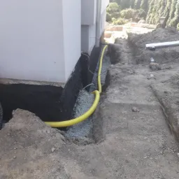 Fundament budynku zabezpieczony czarną izolacją, obsypany żwirem, z ż&oacute;łtą rurą drenażową ułożoną w wykopie wzdłuż ściany.