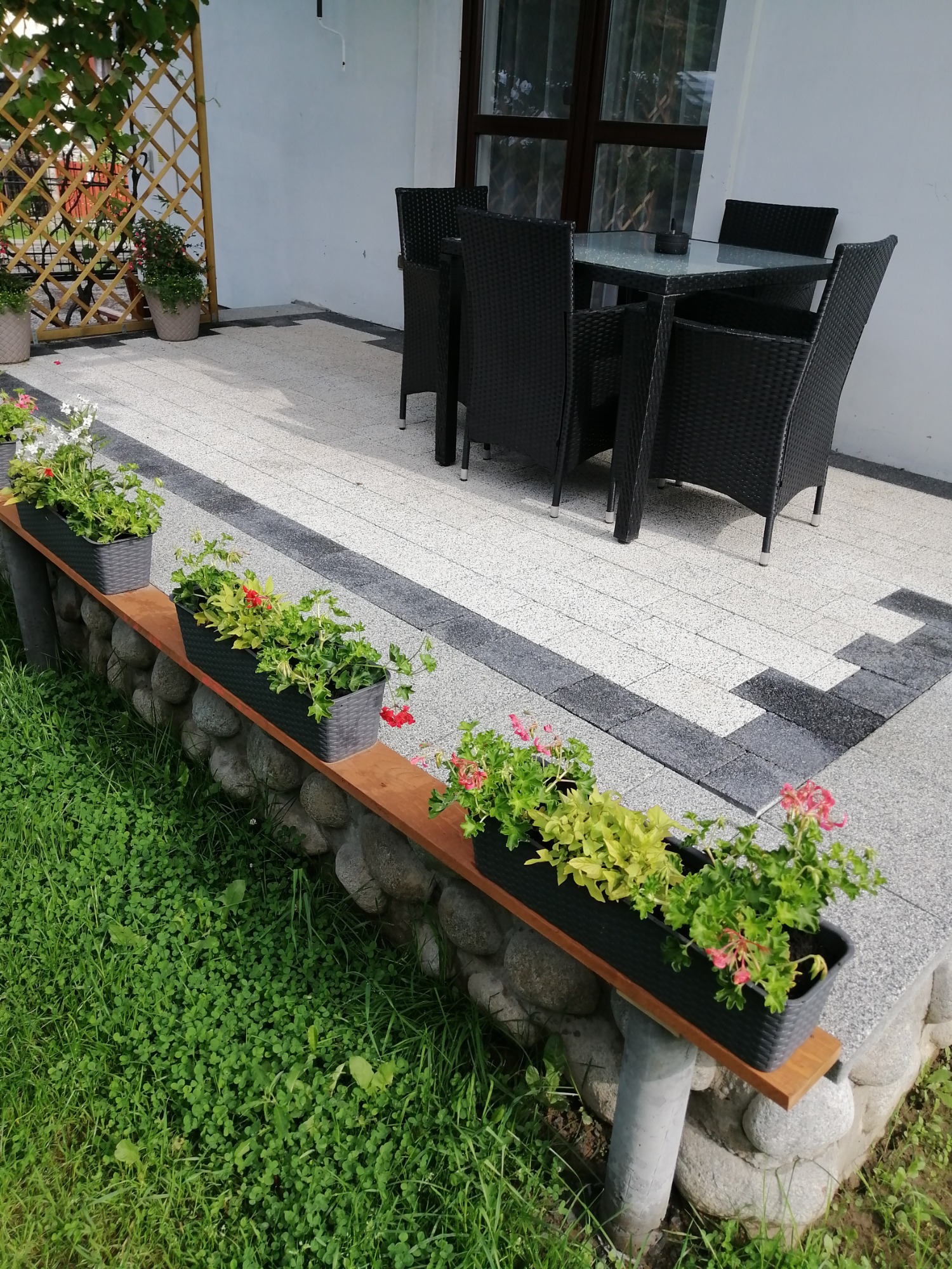 Taras z jasnoszarej kostki brukowej z ciemnoszarym obramowaniem, cztery czarne krzesła i szklany stół na zewnątrz, donice z kwiatami wzdłuż krawędzi tarasu.
