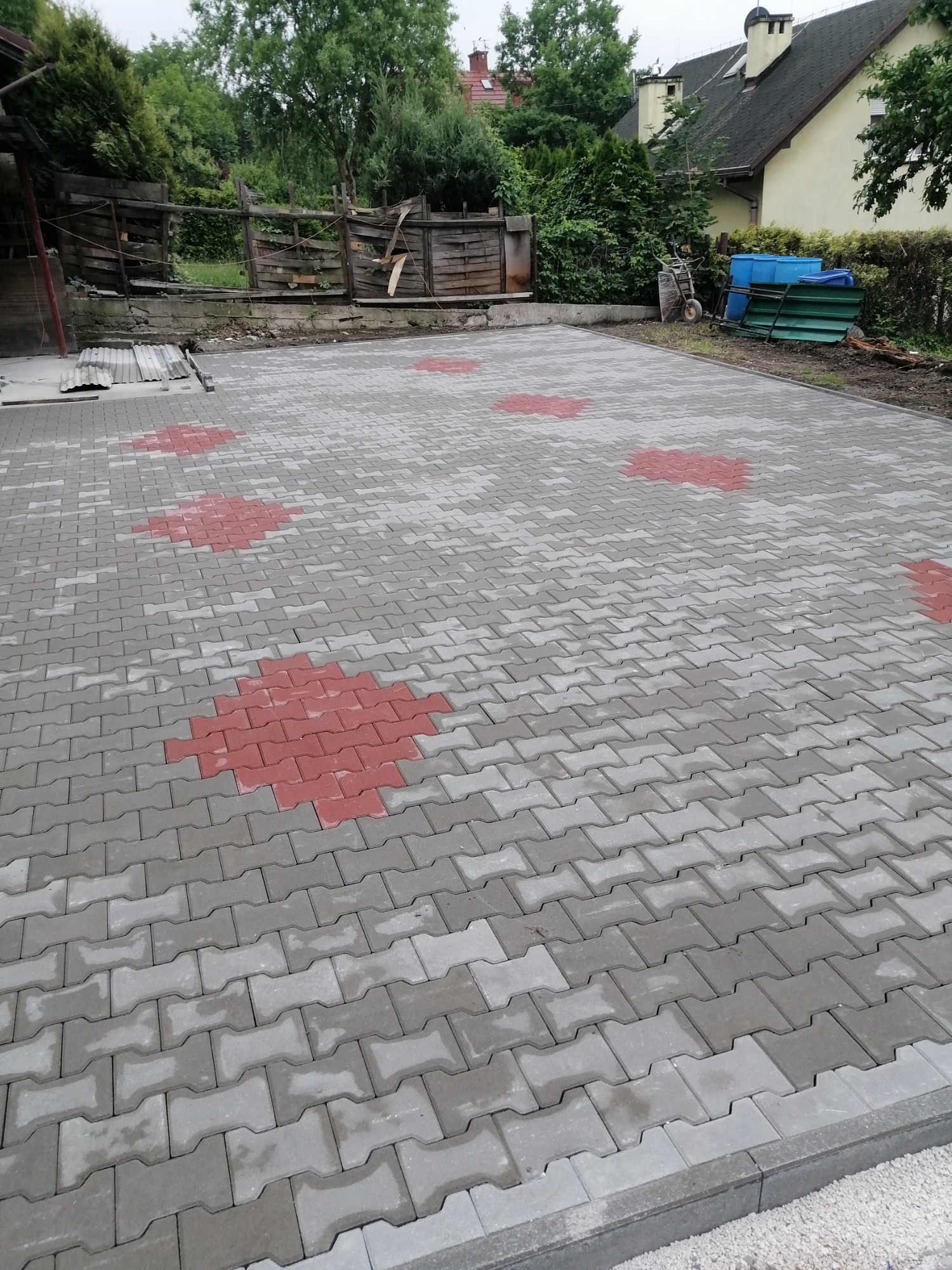 Ułożona nawierzchnia z kostki brukowej w odcieniach szarości, z dekoracyjnymi wstawkami z czerwonej kostki w kształcie rombów, otoczona zielenią i elementami infrastruktury.