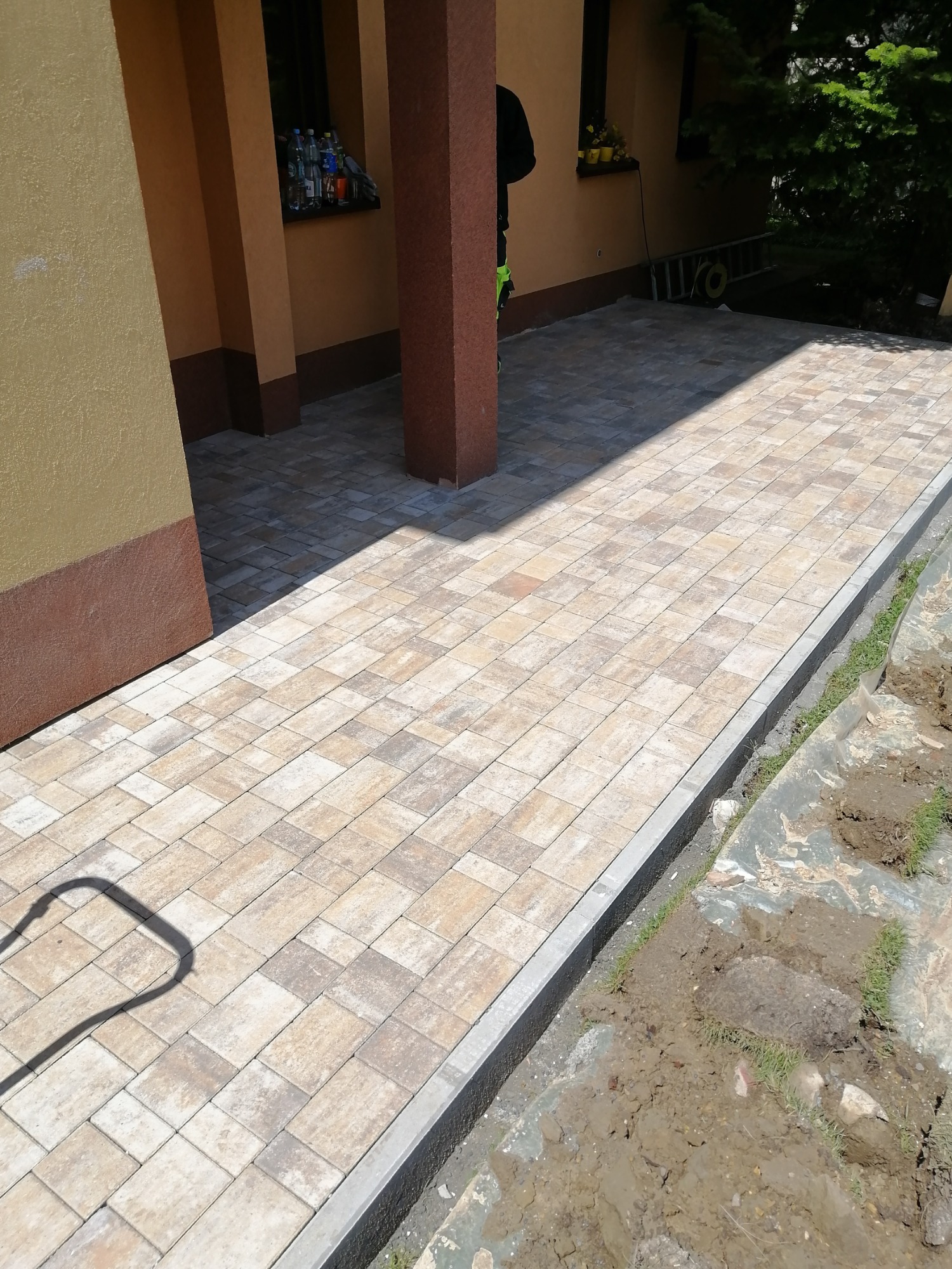 Ułożona kostka brukowa w odcieniach beżu i szarości, z szarym obrzeżem, prowadząca wzdłuż ściany budynku z widocznymi elementami instalacji i fragmentem zieleni z folią ochronną.