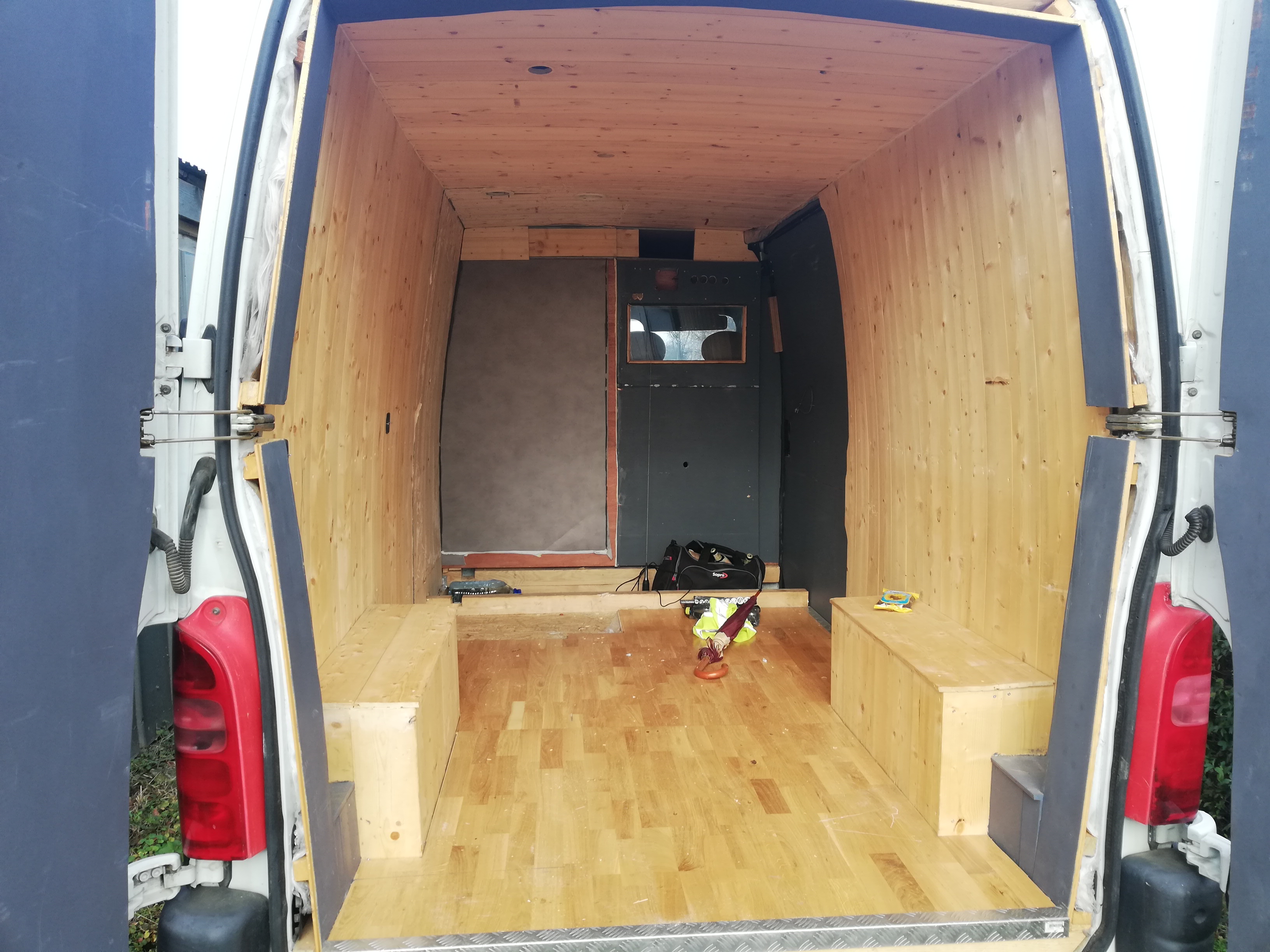 Wnętrze furgonetki w trakcie adaptacji z drewnianą boazerią na ścianach i suficie, częściowo wykończone drewniane zabudowy wzdłuż ścian, widoczne narzędzia i akcesoria robocze.