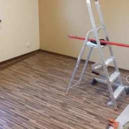 Pokój z ułożonymi panelami podłogowymi w odcieniach brązu, drabina aluminiowa z poziomicą, wiadro z zaprawą i narzędzia do układania paneli.
