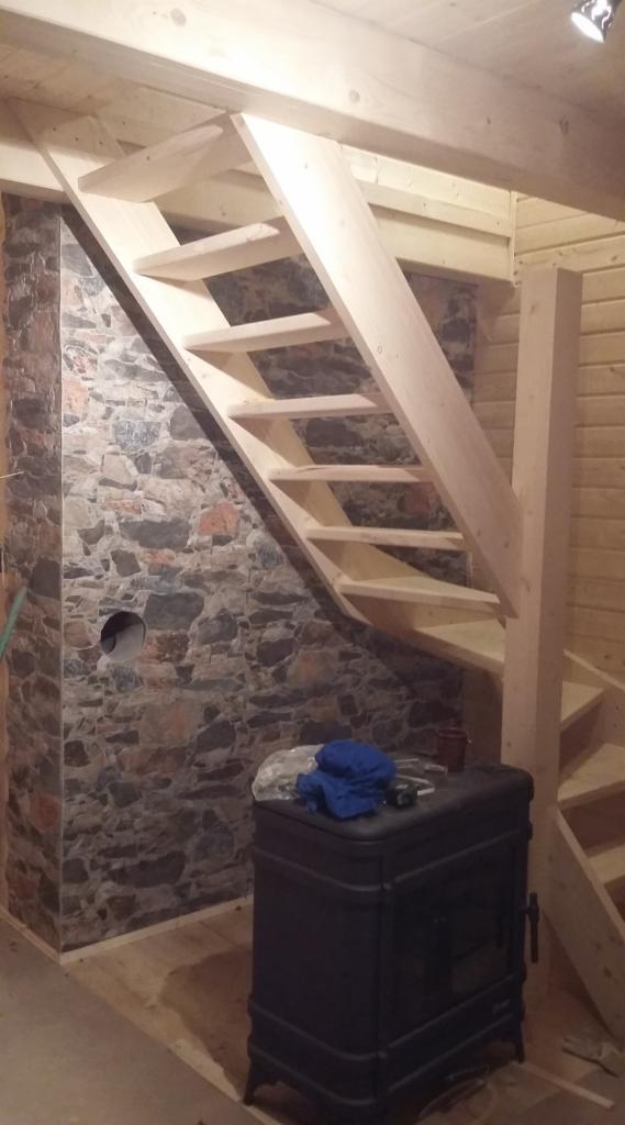 Wnętrze z drewnianymi schodami o surowym wykończeniu, ściana wyłożona imitacją kamienia z otworem, obok piec żeliwny z niebieską szmatką na wierzchu.