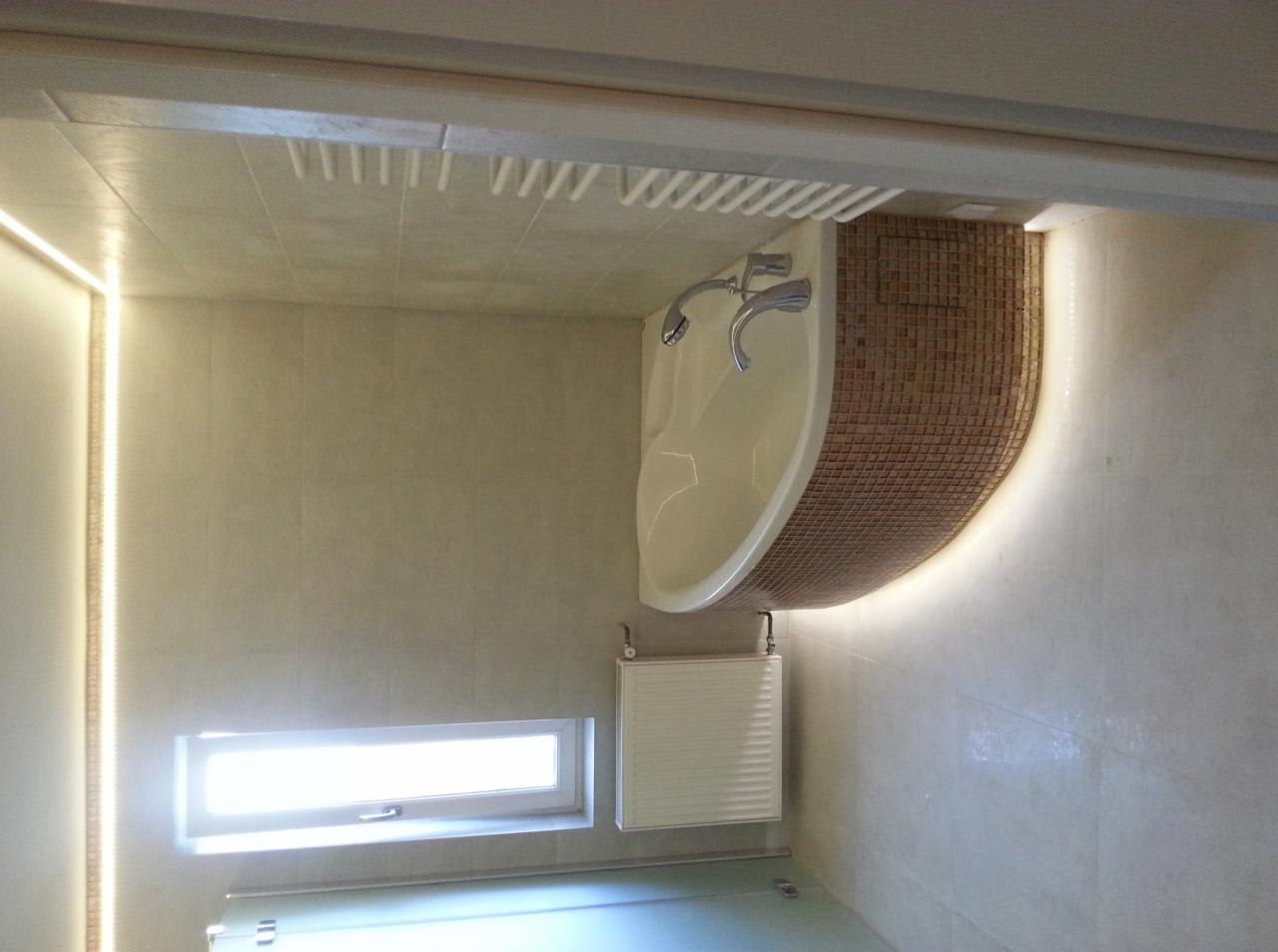 Wykończona łazienka z narożną wanną obudowaną mozaiką, oświetleniem LED i grzejnikiem pod oknem.