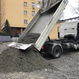 Wyładowywanie betonu z wywrotki na placu budowy, widoczny budynek w tle.