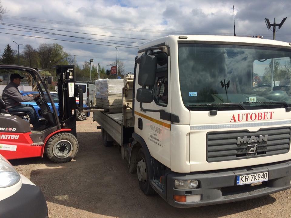 Czerwony wózek widłowy Manitou ładuje paletę z materiałami budowlanymi na białą ciężarówkę MAN z napisem AUTOBET, widoczne tablice rejestracyjne KR 7K940.