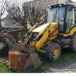 Żółta koparko-ładowarka JCB z łyżką i osprzętem do wykopów, zaparkowana na trawniku obok kostki brukowej i drewnianego płotu. Widoczny element szamba w łyżce.