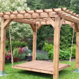 Drewniana pergola z huśtawką-platformą zawieszoną na linach, otoczona zielenią ogrodu z kwitnącymi roślinami, widok na drewniany płot w tle.