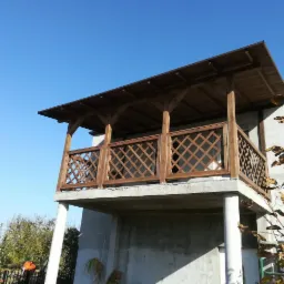 Drewniany balkon z zadaszeniem, wykończony kratownicą, w Gniewkowie. Widok z dołu ukazuje betonową konstrukcję wsporczą i kolumny.