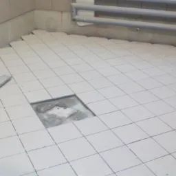 Układanie jasnych, kwadratowych płytek podłogowych z krzyżykami dystansowymi. Brak jednej płytki, widoczny fragment podłoża. Metalowa listwa w lewym rogu.