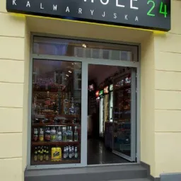 Fasada sklepu z alkoholem z podświetlanym szyldem 'Alkohole Kalwaryjska 24' nad wejściem, widoczne butelki i lodówki wewnątrz.